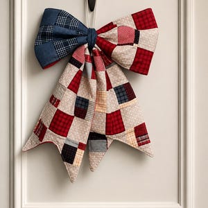 May include: A large decorative bow constructed from a patchwork of fabric squares. The design features a mix of red, navy blue, and beige patterns, including gingham, polka dots, and solid colors. The bow has two long, pointed tails hanging down.