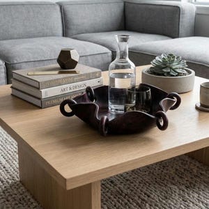 May include: A dark brown, square-shaped decorative bowl with rounded, looped edges. The bowl is crafted with a smooth, curved design and sits on a flat surface. It contains a clear glass carafe filled with water and a small, dark glass tumbler.