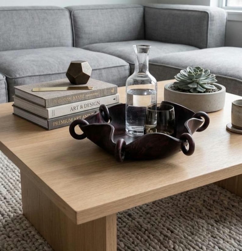 May include: A dark brown, square-shaped decorative bowl with rounded, looped edges. The bowl is crafted with a smooth, curved design and sits on a flat surface. It contains a clear glass carafe filled with water and a small, dark glass tumbler.