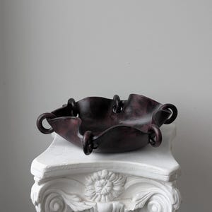 May include: A dark purple and black marbled bowl with a wavy, scalloped edge. The bowl features several circular loops attached along its rim, creating a distinct, interconnected structure. The surface has a smooth, polished appearance throughout.