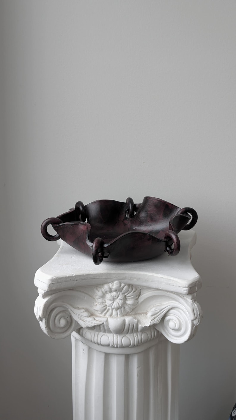 May include: A dark purple and black marbled bowl with a wavy, scalloped edge. The bowl features several circular loops attached along its rim, creating a distinct, interconnected structure. The surface has a smooth, polished appearance throughout.