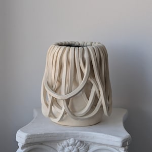 May include: A cream-colored ceramic vase featuring a textured exterior composed of numerous overlapping, curved clay strips that create a cage-like structure around the main body, with a dark interior visible at the top opening of the vessel.