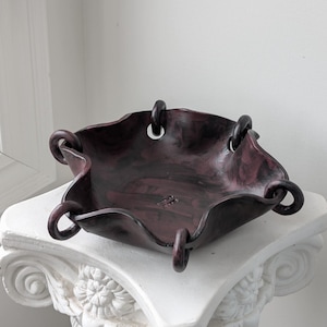 May include: A dark, mottled maroon and black ceramic bowl with a wavy, irregular rim. The bowl features five circular loops attached to the edges, creating a folded, fabric-like appearance. The interior surface displays a subtle, stamped text mark.