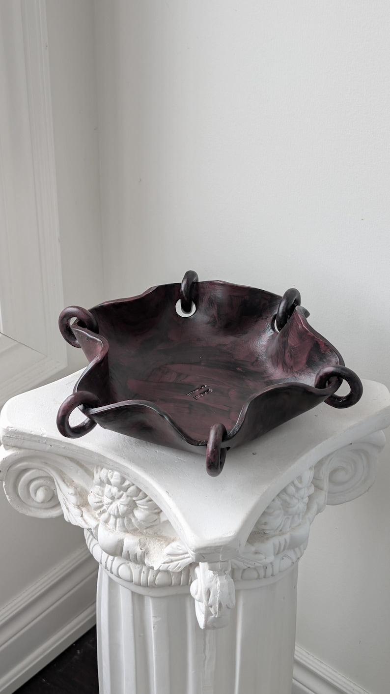 May include: A dark, mottled maroon and black ceramic bowl with a wavy, irregular rim. The bowl features five circular loops attached to the edges, creating a folded, fabric-like appearance. The interior surface displays a subtle, stamped text mark.