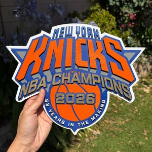 Op de afbeelding: Een meerlaags houten bord met het New York Knicks logo in oranje, blauw, wit en grijs. De tekst luidt New York Knicks, NBA Champions, 2026 en 53 Years in the Making. Het ontwerp bevat een afbeelding van een basketbal.
