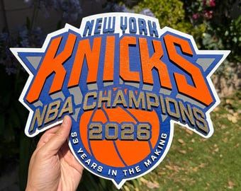 Pode incluir: Uma placa de madeira multicamadas com o logótipo dos New York Knicks em laranja, azul, branco e cinzento. O texto diz New York Knicks, NBA Champions, 2026 e 53 Years in the Making. O design inclui um gráfico de uma bola de basquetebol.