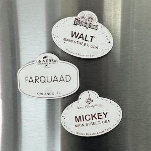 Puede incluir: Tres etiquetas de nombre ovaladas blancas con texto marrón. La etiqueta superior dice "Disneyland" con una pequeña imagen del castillo de Disneyland y dice "Walt, Main Street, USA, Happiest Place on Earth". La etiqueta del medio dice "Universal Orlando Resort" y dice "Farquaad, Orlando, FL". La etiqueta inferior dice "Walt Disney World" y dice "Mickey, Main Street, USA, Where Dreams Come True".
