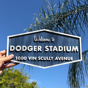 以下が含まれることがあります： 「Welcome to Dodger Stadium 1000 Vin Scully Avenue」と書かれた青と白の看板。看板は、縦線と白い文字が入った青色の背景です。青い空を背景に看板が掲げられています。