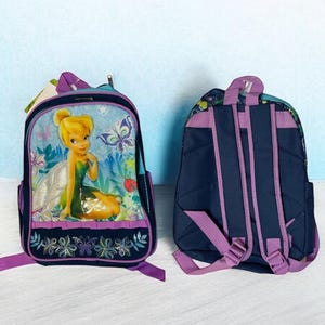 May include: Two children's backpacks. One backpack features a cartoon image of a fairy with a green dress and yellow hair, surrounded by butterflies and flowers. The other backpack is navy blue with purple straps.