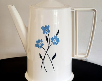 May include: A white plastic coffee pot featuring a central graphic of three blue flowers with dark blue stems and leaves. The pot has a tall cylindrical body, a matching lid with a small knob, a long angled spout, and a rectangular handle.