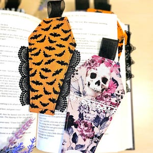 May include: Two coffin-shaped fabric bookmarks with black ribbon tabs. One features an orange background with a black bat pattern and black lace trim. The other displays a skull and floral print in pink and grey tones with white lace trim.