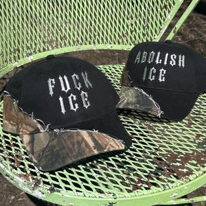 May include: Two black baseball caps with camouflage visors are displayed on a green metal table. One cap has the text "FUCK ICE" embroidered in white, and the other says "ABOLISH ICE". The caps are outdoors.