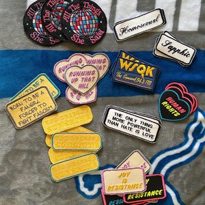 Puede incluir: Un conjunto de parches bordados con varios diseños y texto. Algunos parches son redondos con un diseño de globo y el texto "All The Things She Said". Otros parches tienen frases como "You Deserve Sunshine" y "Human Rights."