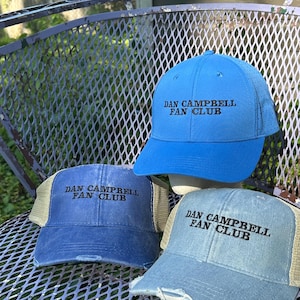 Op de afbeelding: Drie blauwe baseballpetten, elk geborduurd met "DAN CAMPBELL FAN CLUB" in zwart. De hoeden hebben verschillende stijlen, waaronder een truckerpet met een mesh achterkant en een geperforeerde pet. De hoeden worden tentoongesteld op een metalen stoel.