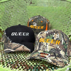 May include: Three baseball caps with the word "QUEER" in white or yellow lettering. One cap is black with a camouflage brim, another is camouflage with an orange brim, and the third is camouflage with a mesh back.