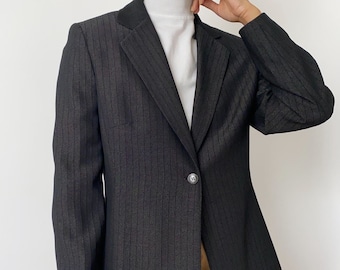May include: A black blazer with a subtle pinstripe pattern. The blazer has a single-button closure and a notched lapel. The model is wearing a white turtleneck and a tan skirt. The blazer is a classic piece of clothing.