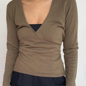 May include: Olive green wrap-style long-sleeve top. The top features a deep V-neck and a fitted silhouette. The sleeves are long and the fabric appears to be a knit material. The top is paired with black pants. The background is a plain white wall.