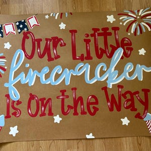 May include: A brown poster with the text "Our Little Firecracker is On the Way" in red and light blue. The poster features illustrations of fireworks, stars, and patriotic bunting.