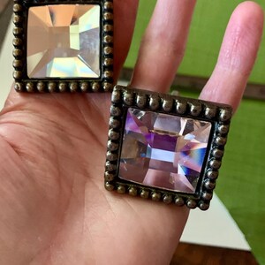 May include: Two square cabinet knobs with a bronze finish and large, faceted, clear crystal centres. Each knob has a beaded border. The crystals reflect light, creating a sparkling effect. The knobs are held in a hand against a green background.
