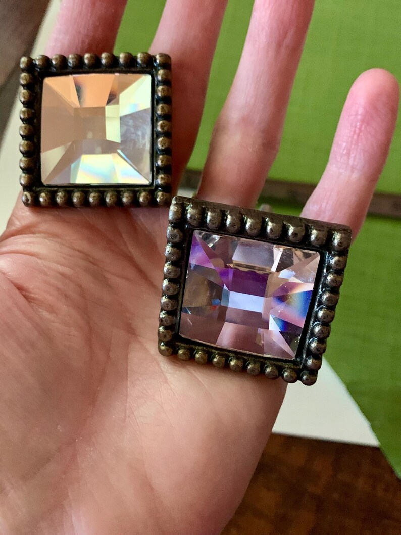 May include: Two square cabinet knobs with a bronze finish and large, faceted, clear crystal centres. Each knob has a beaded border. The crystals reflect light, creating a sparkling effect. The knobs are held in a hand against a green background.