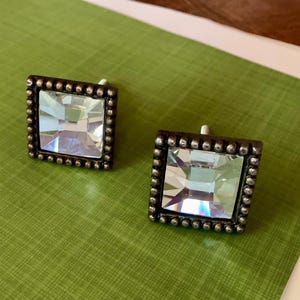 May include: A pair of square earrings featuring a clear, faceted central stone. The earrings are set within a dark metal frame with small, round detailing. They are displayed on a textured green surface.