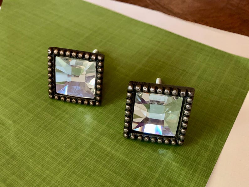 May include: A pair of square earrings featuring a clear, faceted central stone. The earrings are set within a dark metal frame with small, round detailing. They are displayed on a textured green surface.