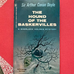 May include: A vintage paperback book cover featuring a spooky illustration of a skull, magnifying glass, pipe, and a detective's hat in a shadowy forest. The book title is "The Hound of the Baskervilles" and the author is "Sir Arthur Conan Doyle".