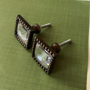 May include: Two cabinet knobs with a decorative design. Each knob has a clear, faceted, rectangular crystal set in a square, dark bronze-coloured frame with a beaded edge. They have a threaded post for mounting.