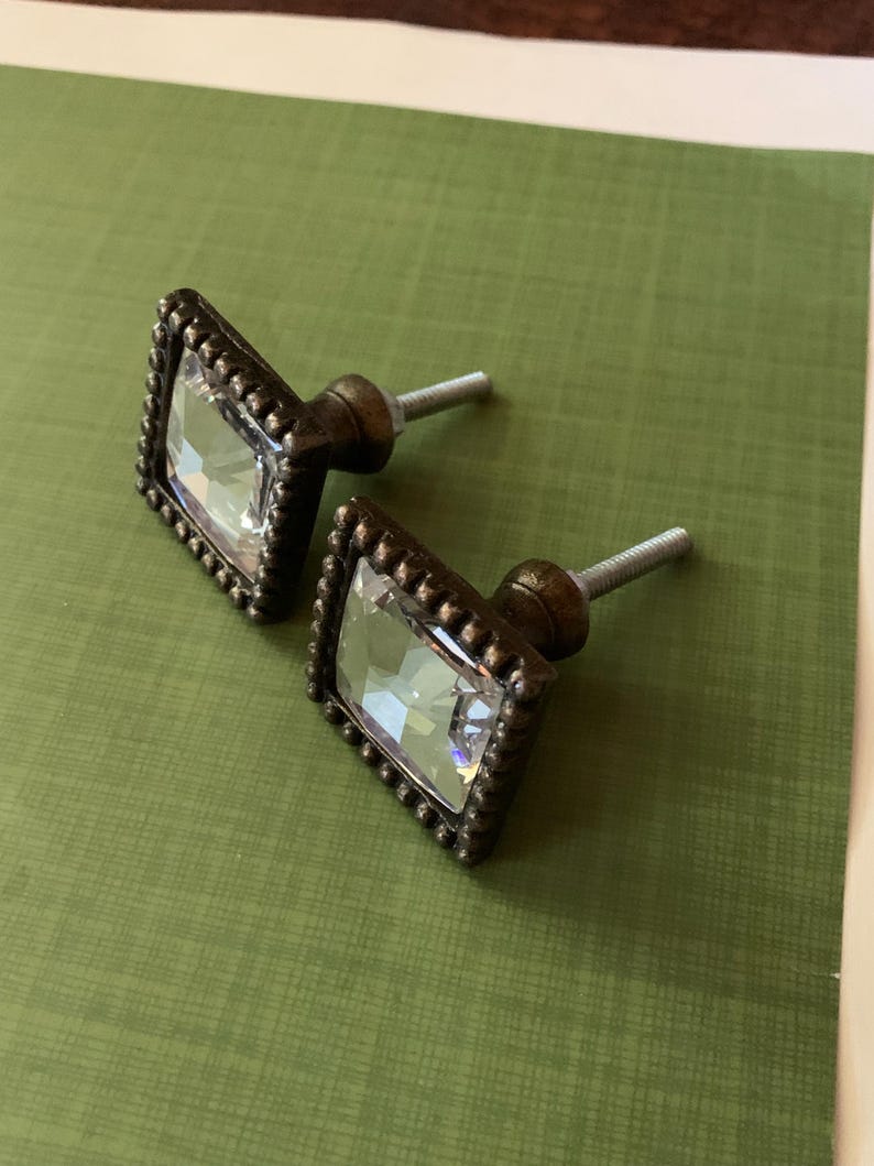 May include: Two cabinet knobs with a decorative design. Each knob has a clear, faceted, rectangular crystal set in a square, dark bronze-coloured frame with a beaded edge. They have a threaded post for mounting.