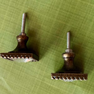 May include: Two bronze-finished cabinet knobs. Each knob features a square base with a beaded edge and a clear, faceted crystal centre. The knobs are set against a green background.