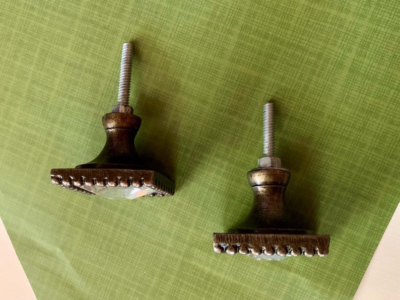 May include: Two bronze-finished cabinet knobs. Each knob features a square base with a beaded edge and a clear, faceted crystal centre. The knobs are set against a green background.