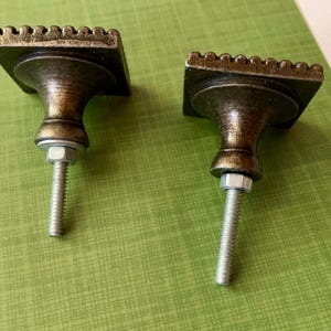 May include: Two antique-style cabinet knobs. Each knob has a square, textured top with a decorative edge, a rounded body, and a threaded post with a nut. The knobs are bronze-coloured and sit on a green surface.