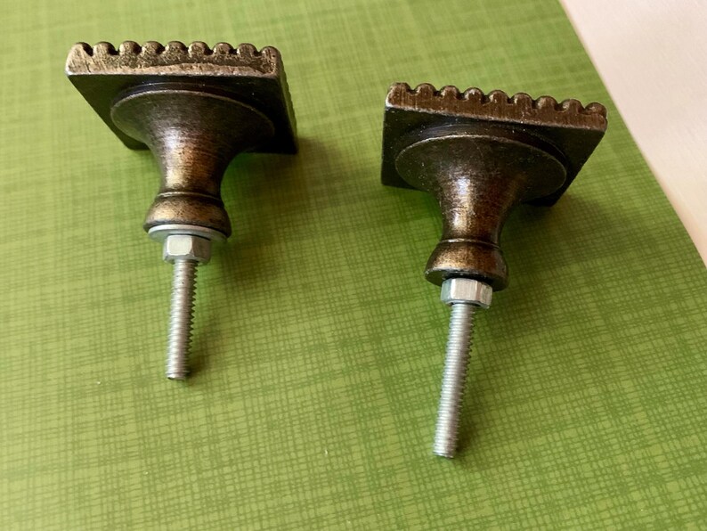 May include: Two antique-style cabinet knobs. Each knob has a square, textured top with a decorative edge, a rounded body, and a threaded post with a nut. The knobs are bronze-coloured and sit on a green surface.