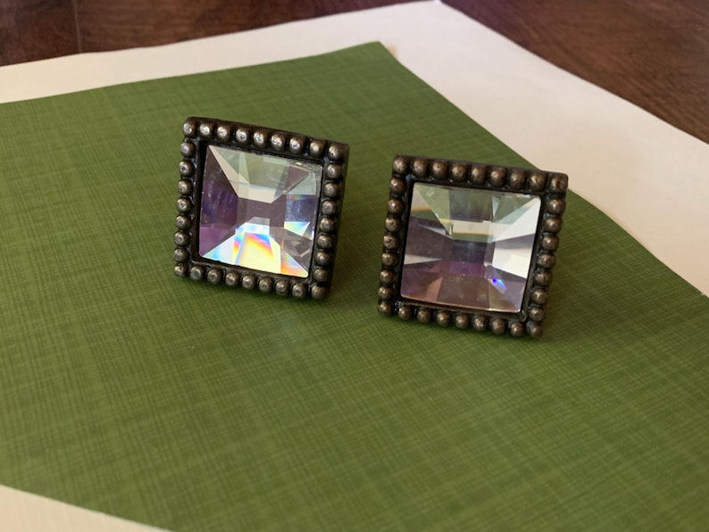 May include: A pair of square stud earrings. Each earring has a large, faceted, clear crystal centre stone with iridescent colour flashes, set in a dark metal frame with a beaded border. The earrings are displayed on a green surface.