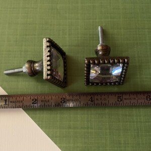 May include: Two decorative cabinet knobs with a square, faceted crystal center and a bronze-coloured metal frame. The knobs are approximately 3.8 cm wide. The knobs are on a green and white surface.