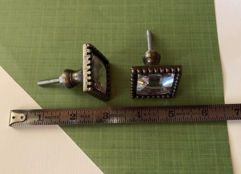 May include: Two decorative cabinet knobs with a square, faceted crystal center and a bronze-coloured metal frame. The knobs are approximately 3.8 cm wide. The knobs are on a green and white surface.