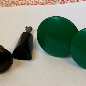 May include: A collection of vintage cabinet knobs and drawer pulls. The knobs are black and round, while the pulls are green and circular. The collection includes a black, angled pull with a silver screw.