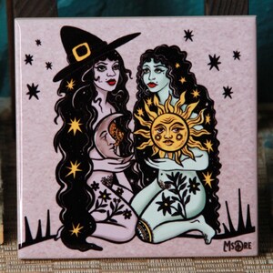 May include: Square ceramic tile with a mystical illustration of two women. One wears a witch's hat and holds a crescent moon, the other holds a sun. The artwork is in black, gold, and pale pink.