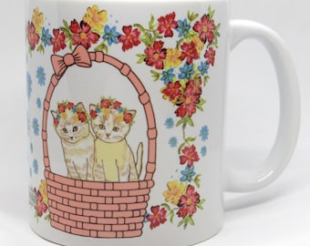 May include: White ceramic mug with a floral design featuring two kittens wearing flower crowns sitting in a pink wicker basket. The mug has a white handle.