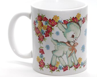 May include: White ceramic mug with a floral design and a cartoon image of a light blue fawn with a pink bow. The fawn is surrounded by pink, yellow, and blue flowers.
