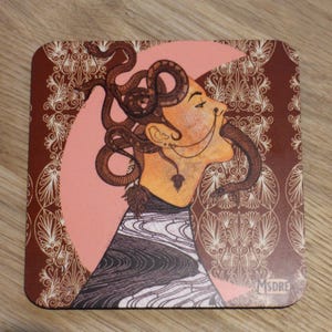 Puede incluir: Un posavasos cuadrado con fondo marrón y un patrón blanco y dorado. El centro del posavasos presenta un círculo rosa con el rostro de una mujer y una serpiente envuelta alrededor de su cabeza. El rostro de la mujer mira hacia la derecha y su boca está abierta. La serpiente es marrón y negra y tiene un patrón blanco y dorado. El texto "MSDRE" está en la esquina inferior derecha del posavasos.