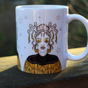 May include: White ceramic mug with a black and gold illustration of a person with long hair and a crown. The background is white with stars.