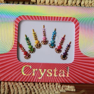 May include: A pack of crystal forehead stickers in a rainbow of colours. The stickers are arranged in a spectrum, from red to purple. The package has a red frame with the word "Crystal" printed on it.