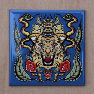May include: A blue square magnet featuring a yellow and orange tiger face with a third eye on its forehead. The tiger is surrounded by two snakes with red eyes and green leaves. The text "MSDRE" is above the tiger's head.