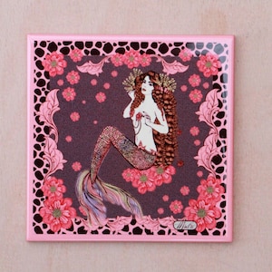 May include: A pink square ceramic tile with a brown and pink floral design. A mermaid with long brown hair and a pink tail sits on a bed of pink flowers. The tile has a pink border and a black and white leopard print pattern.