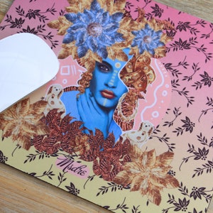 May include: A blue and gold mouse mat with a floral design and a woman's face in the centre. The woman has blue skin and is wearing a crown of flowers. The text "Misdre" is written in the bottom right corner.