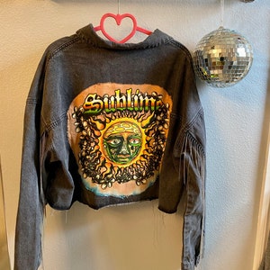 May include: A black denim jacket with a distressed hem and fringe detailing on the sleeves. The back of the jacket features a colorful patch with the word "Sublime" and a graphic of a sun with a face.