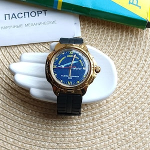 May include: A gold-tone wristwatch with a black band. The watch face is blue with white lettering and a graphic of a bridge and a bird. The watch face reads "Dnipropetrovsk 2000" and "Паден Бого Мосту".