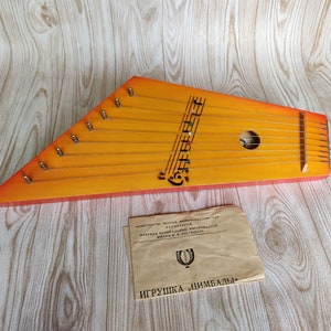 Puede incluir: Un dulcimer de madera naranja y rojo con cuerdas y un agujero de sonido. El dulcimer tiene un diseño tallado de notas musicales y la palabra "Цимбалы" en la parte delantera. El dulcimer está sobre una superficie de madera con una hoja de instrucciones de papel que dice "Игрушка "Цимбалы" en ruso.