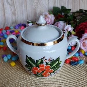 May include: White porcelain sugar bowl with a gold trim and a floral design featuring red berries and green leaves. The lid has a pointed top.